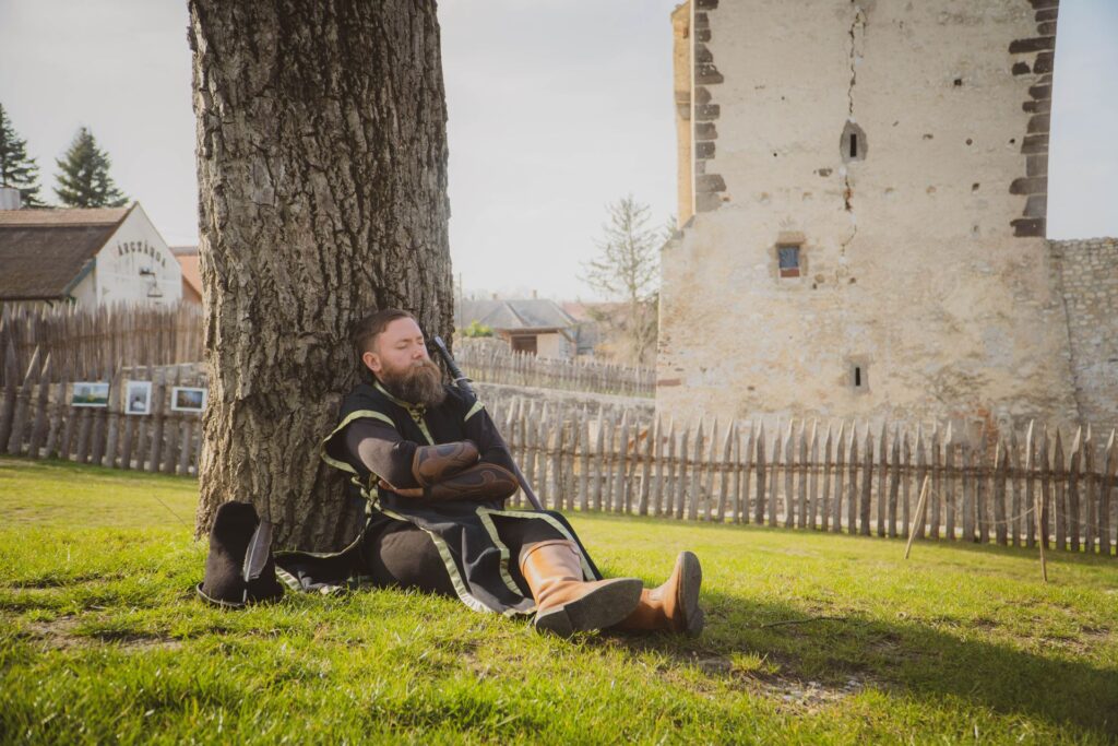 Középkori öltözetű, szakállas férfi pihen a fa tövében, háttérben a vár tornyával.